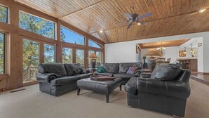 A spacious living room with leather sofas, a coffee table, and a ceiling fan. Large windows offer views of the outdoors. - Country Club Home with a Hot Tub (Flagstaff)