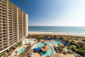 A high-rise hotel with multiple balconies overlooking a beach and a pool area with sun loungers and umbrellas. - Beachfront 2BR Condo with Balcony & Jetted Tub (North Myrtle Beach)