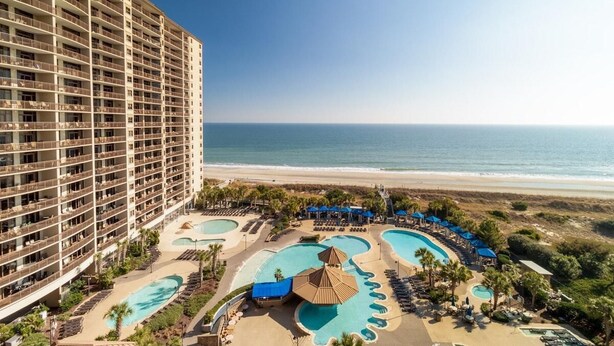 Un hôtel de grande hauteur doté de nombreux balcons donnant sur une plage et une piscine, avec des chaises longues et des parasols.