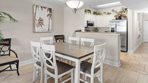 Un coin repas avec une table en bois et des chaises blanches, une cuisine avec des meubles blancs et des appareils en acier inoxydable, et un luminaire suspendu.