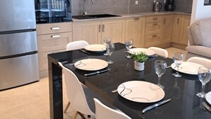 A modern kitchen with wooden cabinets, a black countertop, and a dining area set for six with white chairs and plates. - Gite des Falaises de Longues 6 p (Longues-sur-Mer)