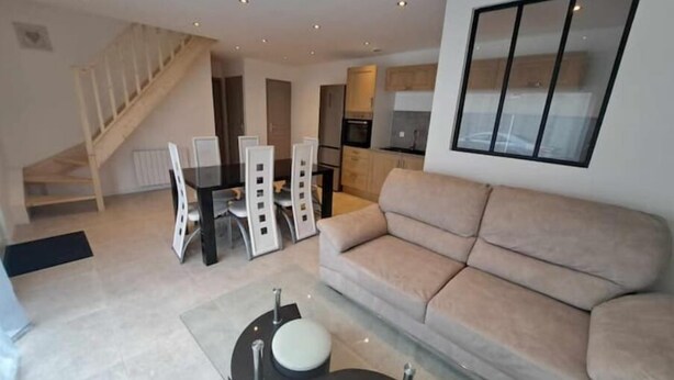 A modern living room with a beige sofa, a dining table with chairs, a staircase, and a kitchen area. - Gite des Falaises de Longues 6 p (Longues-sur-Mer)