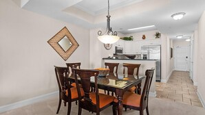 Un comedor con mesa de vidrio, seis sillas de madera con cojines naranja, una lámpara colgante y una cocina al fondo.