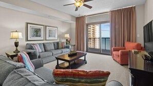 Una sala de estar con un sofá gris, un sillón rojo, una mesa de centro y un televisor. Hay un balcón con cortinas y vistas a edificios.