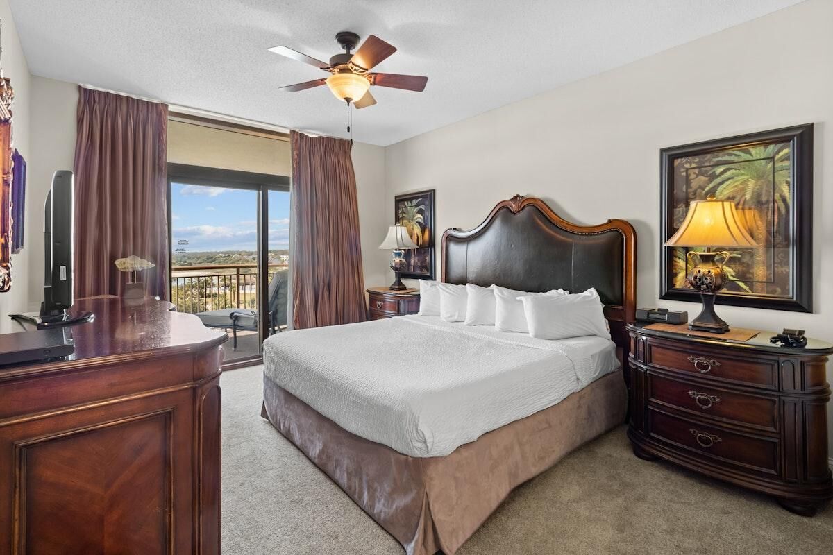 Habitación de hotel con una cama grande, ventilador de techo, escritorio con lámpara, cómoda y un amplio ventanal con cortinas.