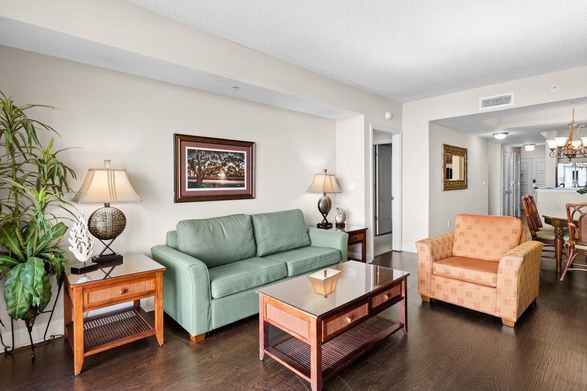 Una sala de estar con un sofá verde, una mesa de centro de madera y un sillón naranja. Hay dos lámparas sobre mesas auxiliares, un cuadro en la pared y una planta grande.