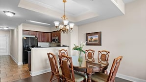 A dining area with a round table, six wooden chairs, a chandelier, and a kitchen in the background. - Sunset Symphony Luxurious 2BR/2BA Oceanfront Condo (North Myrtle Beach)