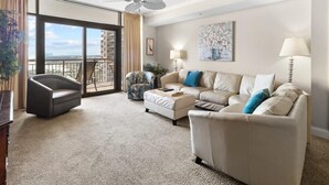 A spacious living room with a large sofa, a chair, and a small table. There is a balcony with a view of the city.