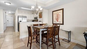 A dining area with a table and chairs, a chandelier, a kitchen in the background, and a framed artwork on the wall.