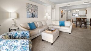 A living room with a sectional sofa, a matching ottoman, and a floral-patterned chair. There is a painting on the wall, a ceiling fan, and a view into the kitchen area.