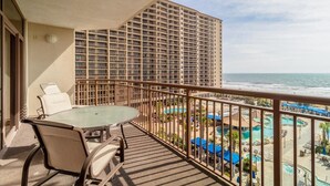 A balcony with a glass table and chairs, overlooking a pool area and a high-rise building. - Wave Watcher's Paradise Cove: 2BR Condo (North Myrtle Beach)