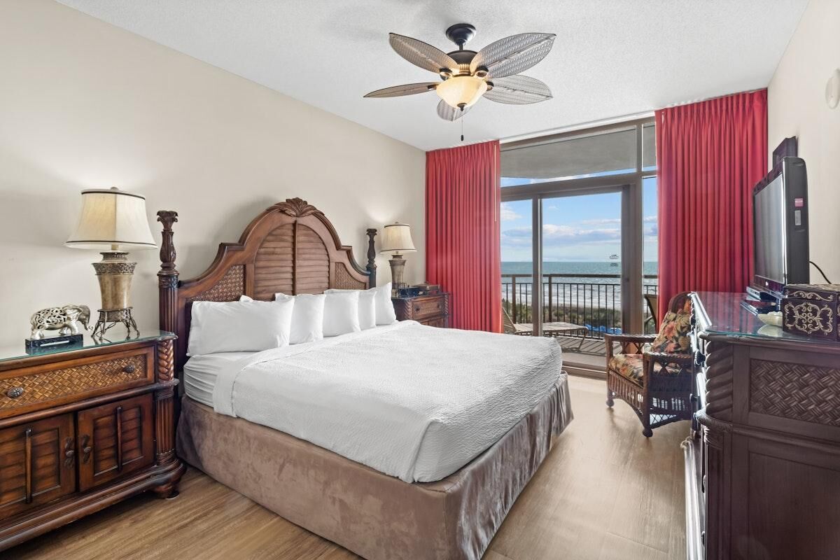 A hotel room with a large bed, two bedside tables with lamps, a ceiling fan, a large window with red curtains, and a view of the ocean.
