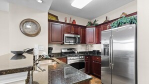 A kitchen with dark wooden cabinets, stainless steel appliances, a granite countertop, and a wall clock. - Wave Watcher's Paradise Cove: 2BR Condo (North Myrtle Beach)