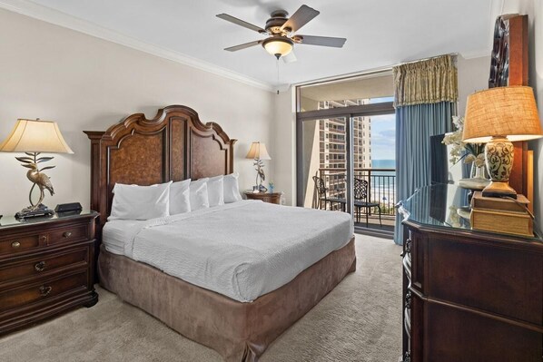 Une chambre d’hôtel avec un grand lit, une tête de lit en bois, deux tables de chevet avec des lampes, un ventilateur de plafond et une vue sur un balcon avec des rideaux bleus.