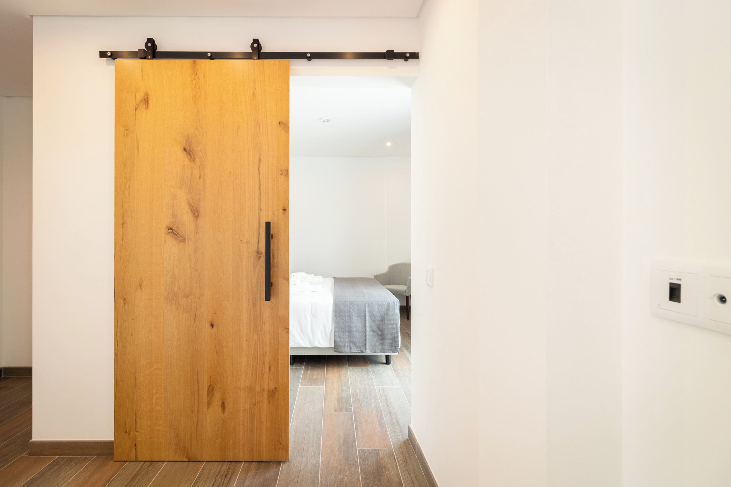 Una habitación con puerta corredera de madera, cama con manta gris y pared blanca con toma de corriente.