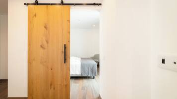 A room with a wooden sliding door, a bed with a gray blanket, and a white wall with a power outlet.