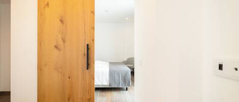 Una habitación con puerta corredera de madera, cama con manta gris y pared blanca con toma de corriente.