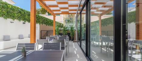 Un patio exterior con mesas y sillas de madera, un muro de cristal y una estructura de pérgola.