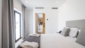 A modern hotel room with a large bed, a bedside table, a wardrobe, and a bathroom visible through an open door.