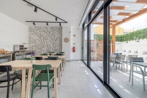 A modern hotel lobby with a long reception desk, green and black chairs, and a large window offering a view of the outdoor seating area.