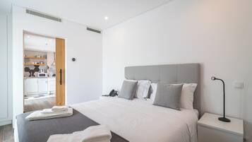 Habitación de hotel moderna con una cama grande, una mesita de noche con lámpara y vista a la cocina por una puerta abierta.