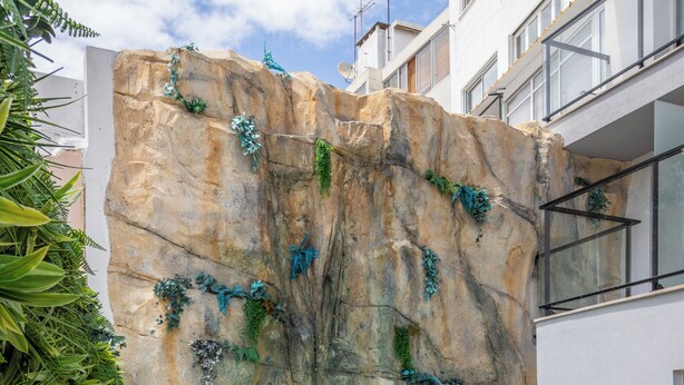A swimming pool with a rock wall, two wicker chairs, and a wooden deck.