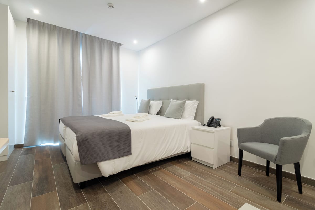 Una habitación de hotel moderna con una cama grande, una mesita de noche con teléfono y un sillón gris.