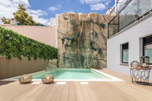 Un área de piscina moderna al aire libre con una pared de roca, dos jarrones decorativos y una mesa de metal con velas.