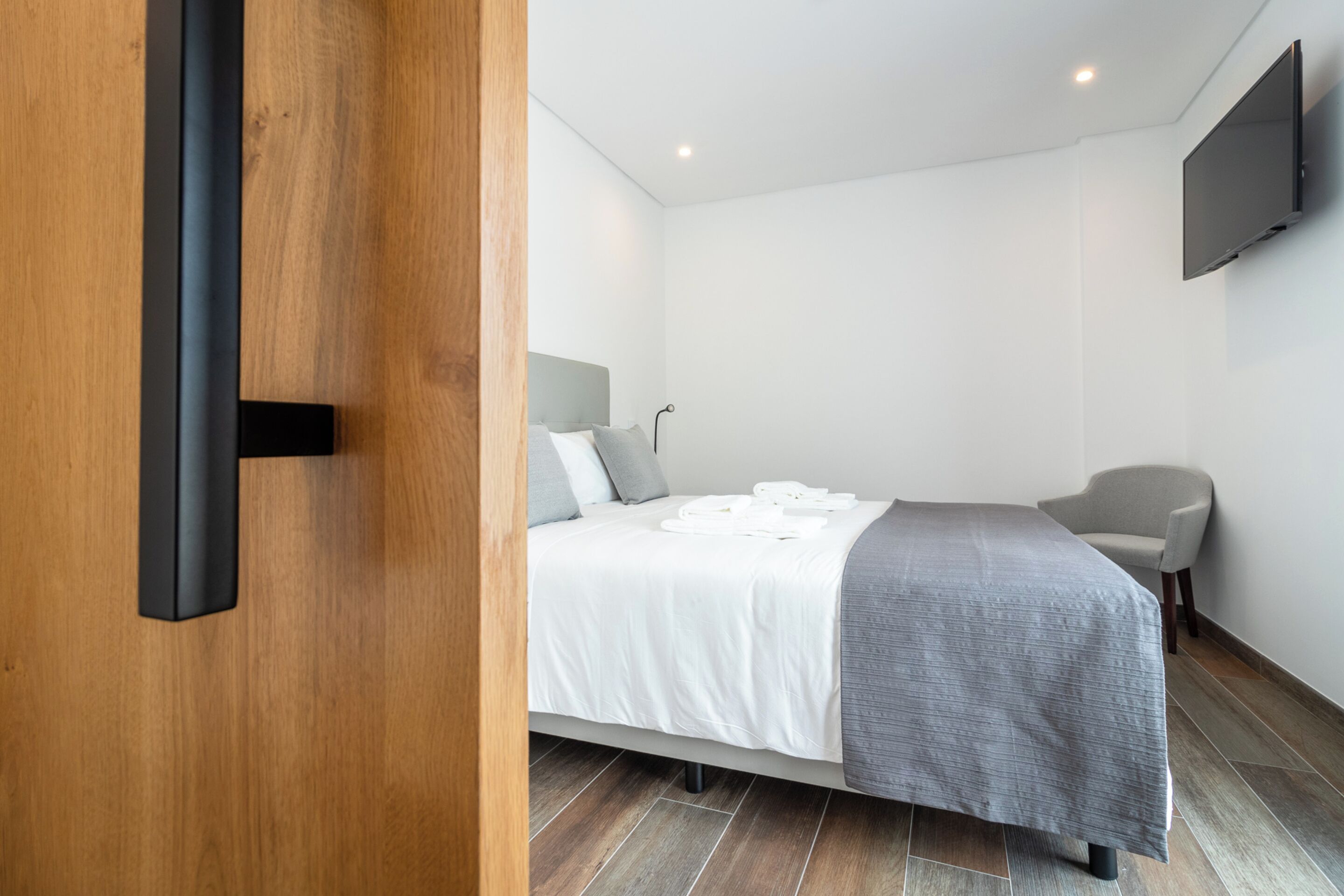 A modern bedroom with a bed, a chair, a flat-screen TV, and a wooden door.