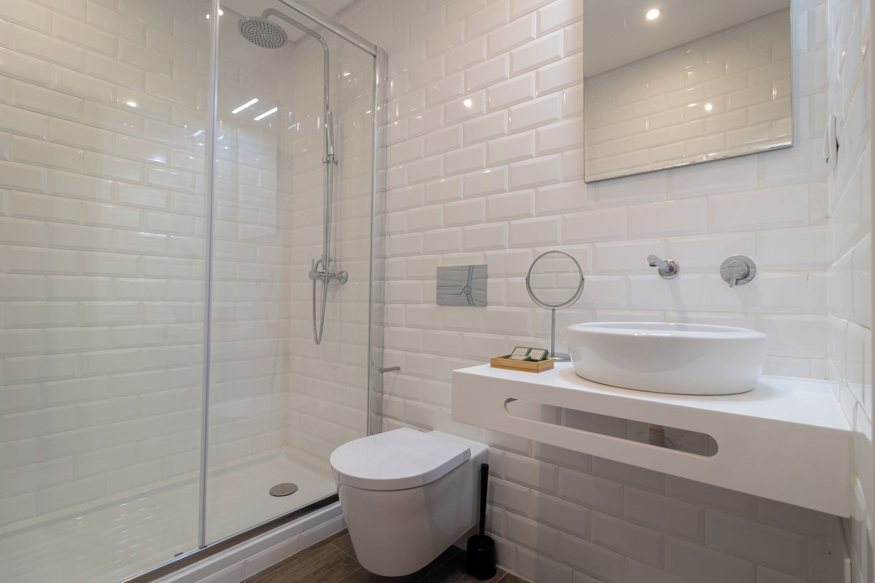 Une salle de bain moderne avec une douche à l’italienne en verre, des murs recouverts de carreaux blancs, un lavabo blanc avec un miroir et des toilettes blanches.