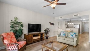 Una sala de estar con televisor, ventilador de techo, sofá, mesa de centro y una cocina al fondo.
