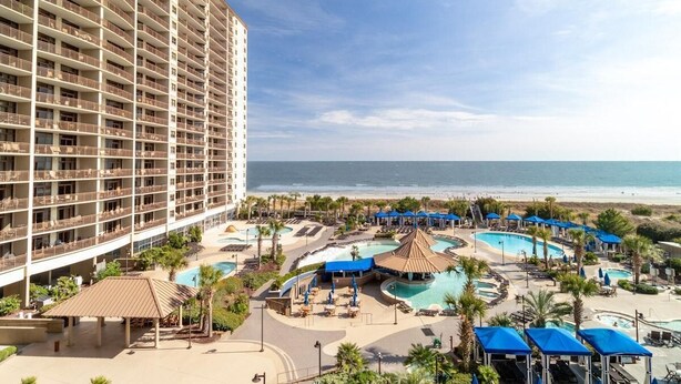 Un hotel con varios balcones, zona de piscina con sombrillas y vistas a la playa.