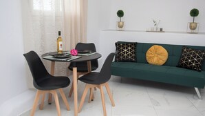 Un comedor con una mesa redonda, cuatro sillas negras, una botella de vino y una flor rosa. Un sofá verde con cojines estampados está apoyado contra la pared.