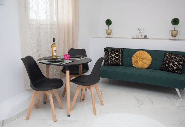Uma área de refeições com uma mesa redonda, quatro cadeiras pretas, uma garrafa de vinho e uma flor rosa. Um sofá verde com almofadas estampadas está encostado na parede.
