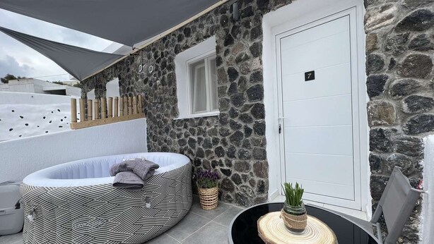Un espacio al aire libre con paredes de piedra, una puerta blanca, una zona de estar circular y una pequeña mesa con una planta.