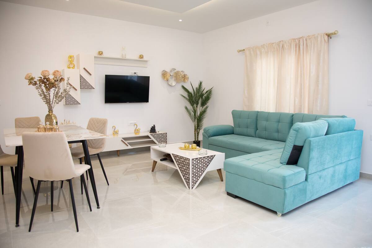 Ein modernes Wohnzimmer mit einem türkisblauen Sofa, einem Esstisch mit Stühlen, einem flachbildfernseher an der Wand und dekorativen Wandbildern.