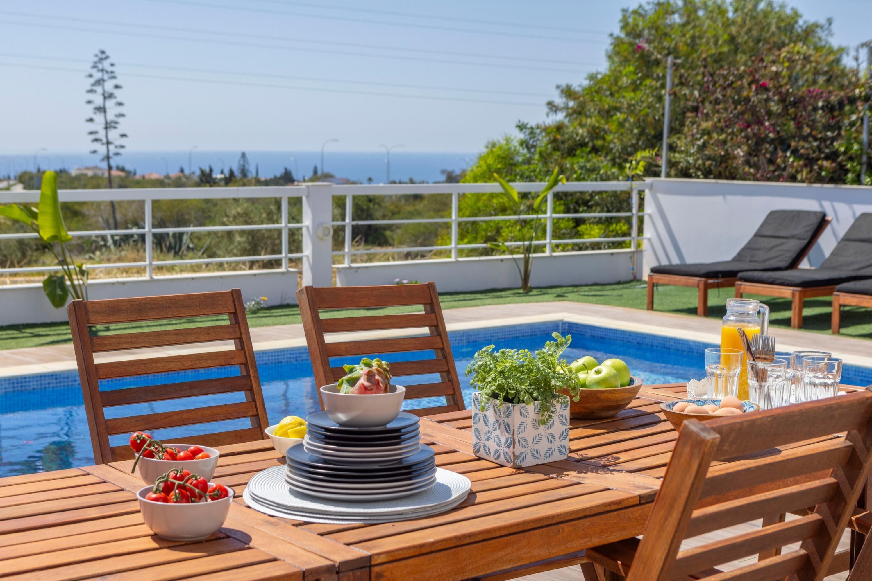 Una mesa de madera con un bol de ensalada, platos y un bol de fruta, cerca de una piscina con sillas de descanso y vistas al mar.