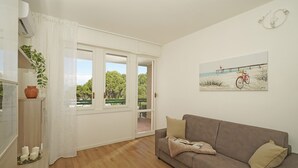 A room with a wooden floor, a brown sofa, a white curtain, a potted plant, and a painting of a bicycle on a beach. - Poggio al Lago 17 by Wonderful Italy (Sirmione)