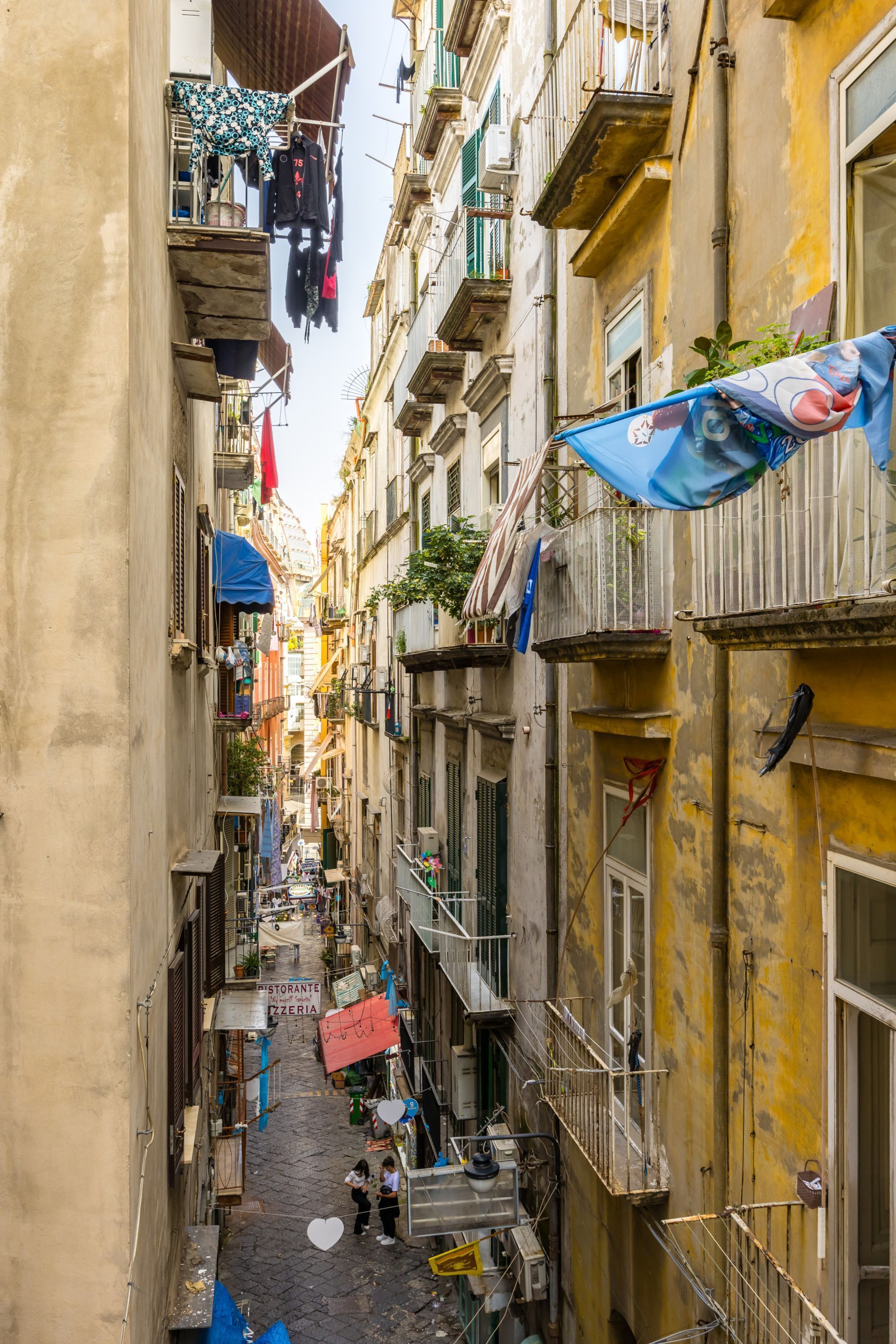Une ruelle étroite entre deux immeubles dotés de balcons, dont certains sont ornés de linge séchant, et une enseigne indiquant un restaurant.