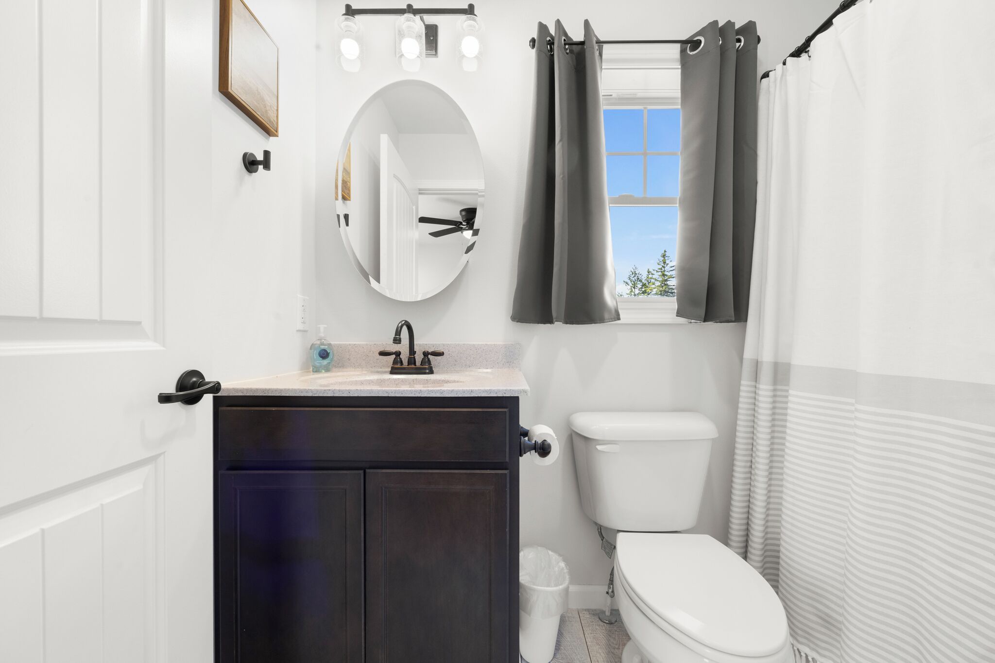 A bathroom with a white toilet, dark cabinets, a round mirror, and a window with curtains.