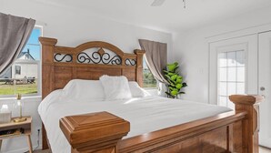 Een slaapkamer met een houten bed, een nachtkastje, een lamp en uitzicht op een huis.