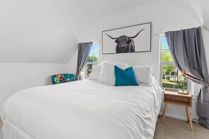 Une chambre à coucher avec un lit, une table de chevet, une lampe, un tableau encadré et une fenêtre avec des rideaux.