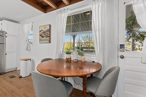 Un coin repas avec une table en bois et quatre chaises, une fenêtre avec des rideaux blancs, un r éfrigérateur et une poubelle.