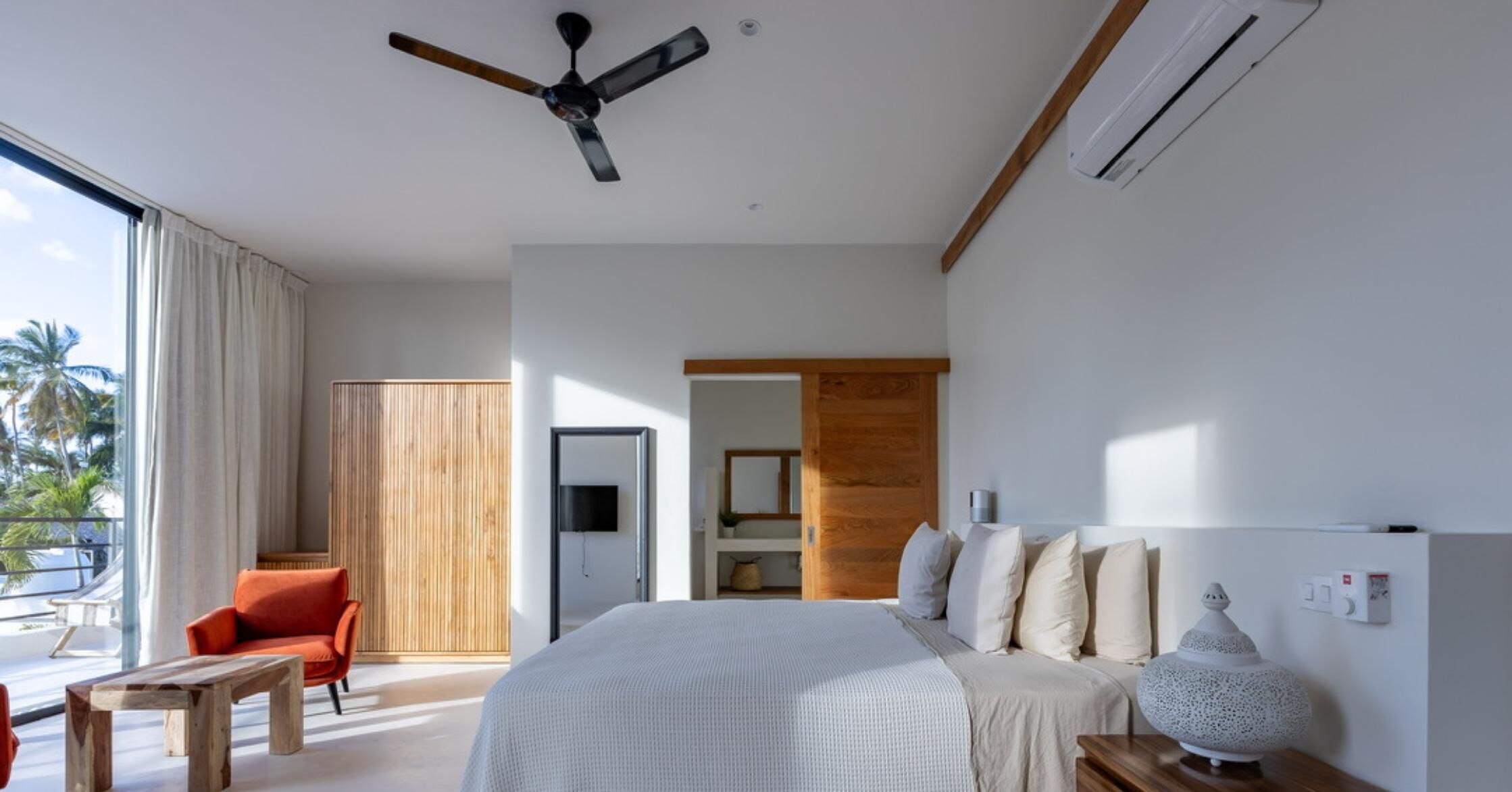 A modern bedroom with a large bed, a ceiling fan, an air conditioning unit, a wooden wardrobe, a red chair, and a view of palm trees through the window.