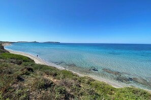 A coastal landscape with clear blue waters, a sandy beach, and green vegetation.