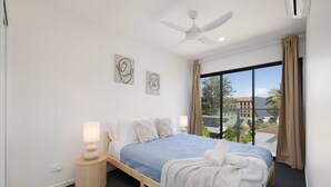 A modern hotel room with a large bed, a ceiling fan, and a view of the city through large windows. - Netflix, Brand new 4BR,Fast NBN,AC, Free Parking (Southport)
