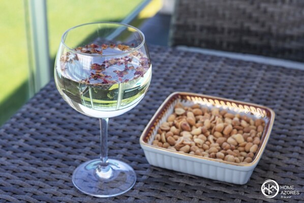 A glass of white wine with dried flowers on the rim, a small dish of almonds, and a wicker table. - Home Azores - Pico do Salomão Poolside Retreat (Ponta Delgada)
