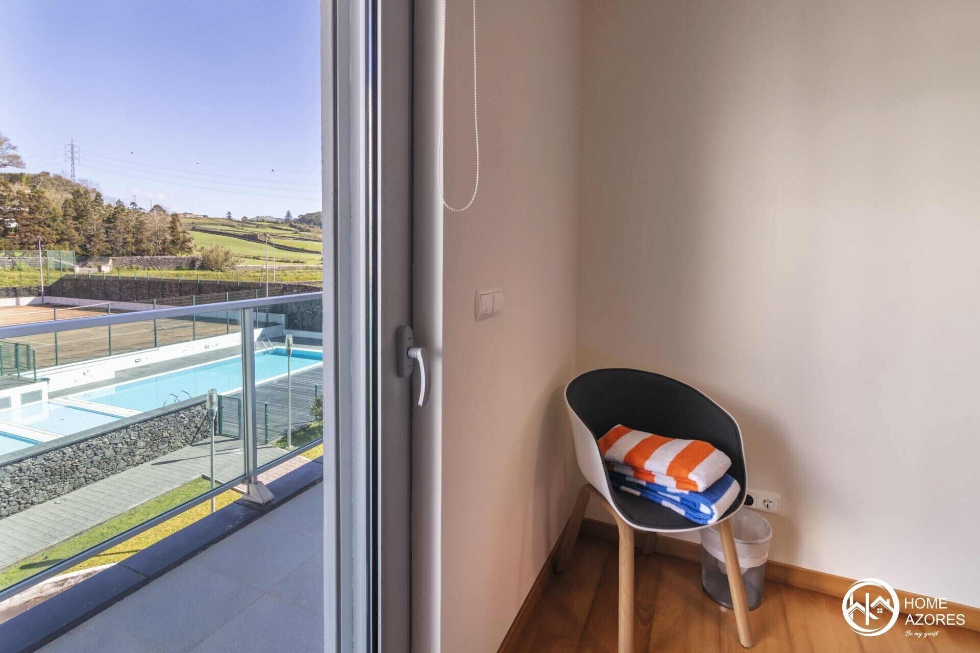 A room with a view of a swimming pool, a chair with towels, and a trash bin.