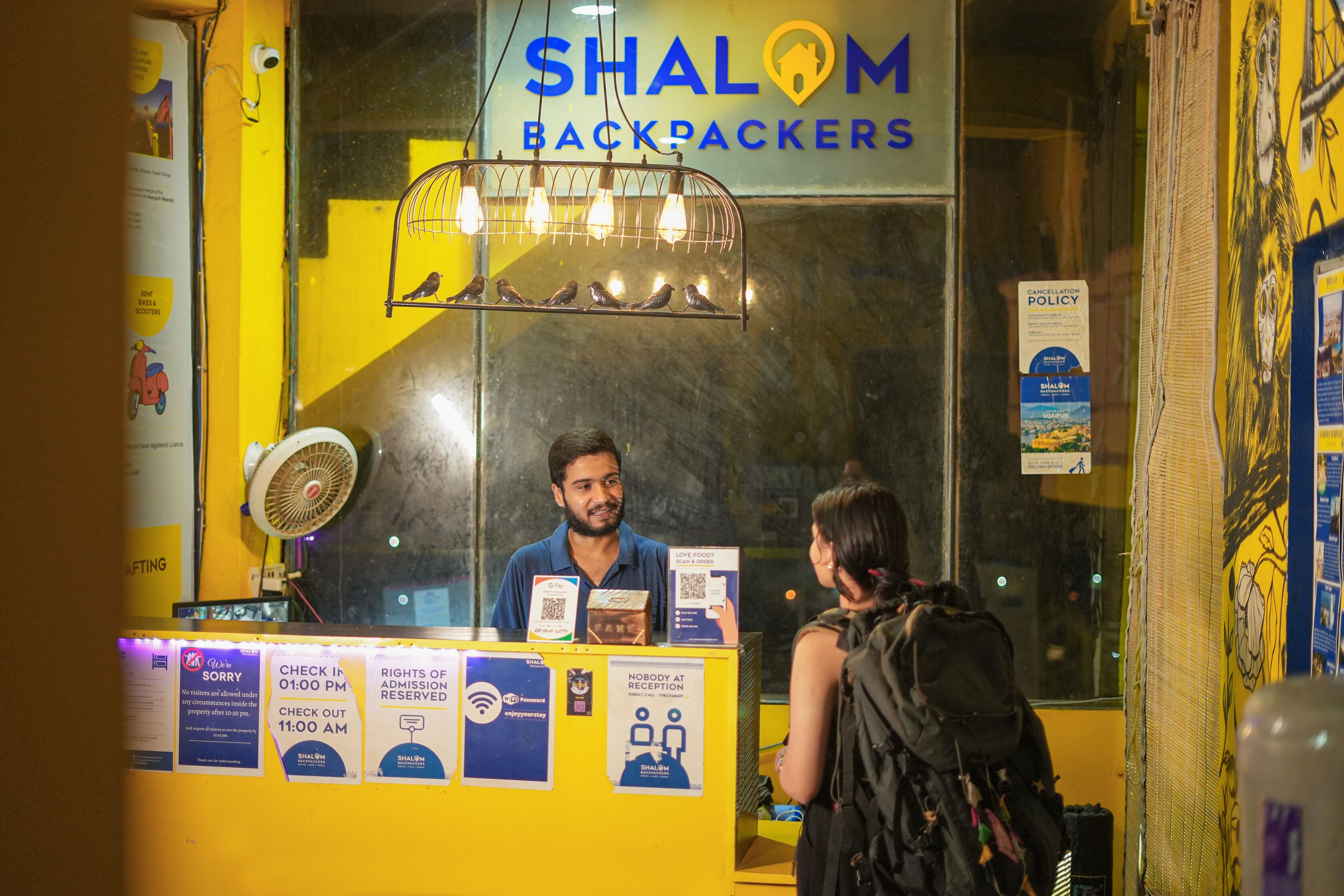 Un comptoir d'accueil chez Shalom Backpackers, avec un panneau, un ventilateur et un code QR pour l'enregistrement.