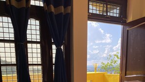 A room with a laptop on a bed, a chair, a table with a book, and a view of a yellow building through a window. - Shalom Backpackers Rishikesh (Narendranagar)
